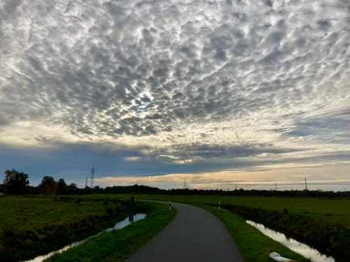 A flat landscape with a paved road, grassy fields and a small canal left and right in front of a cloudy afternoon sky in blues and yellows