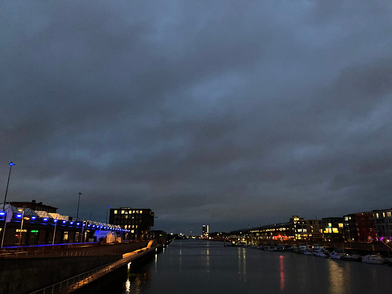 an industrial port in the early morning with dark blue sky and the dark silhouettes of buildings to both sides with lights coming from windows and street lamps etc