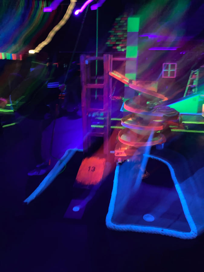 Blurry photo of blacklight minigolf, a bunch of neon colors shining in the dark, the number 13 is readable on a pink background