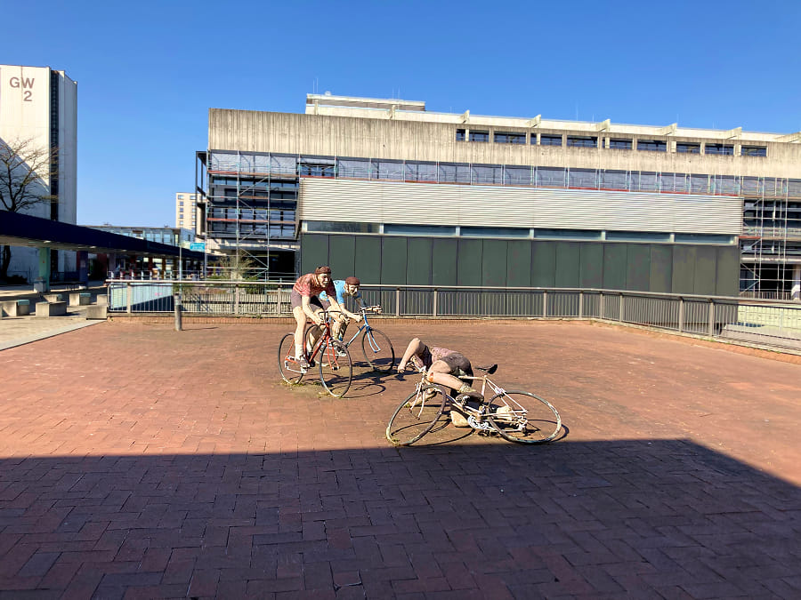 Painted, purous looking sculptures of three road cyclists of which on is falling