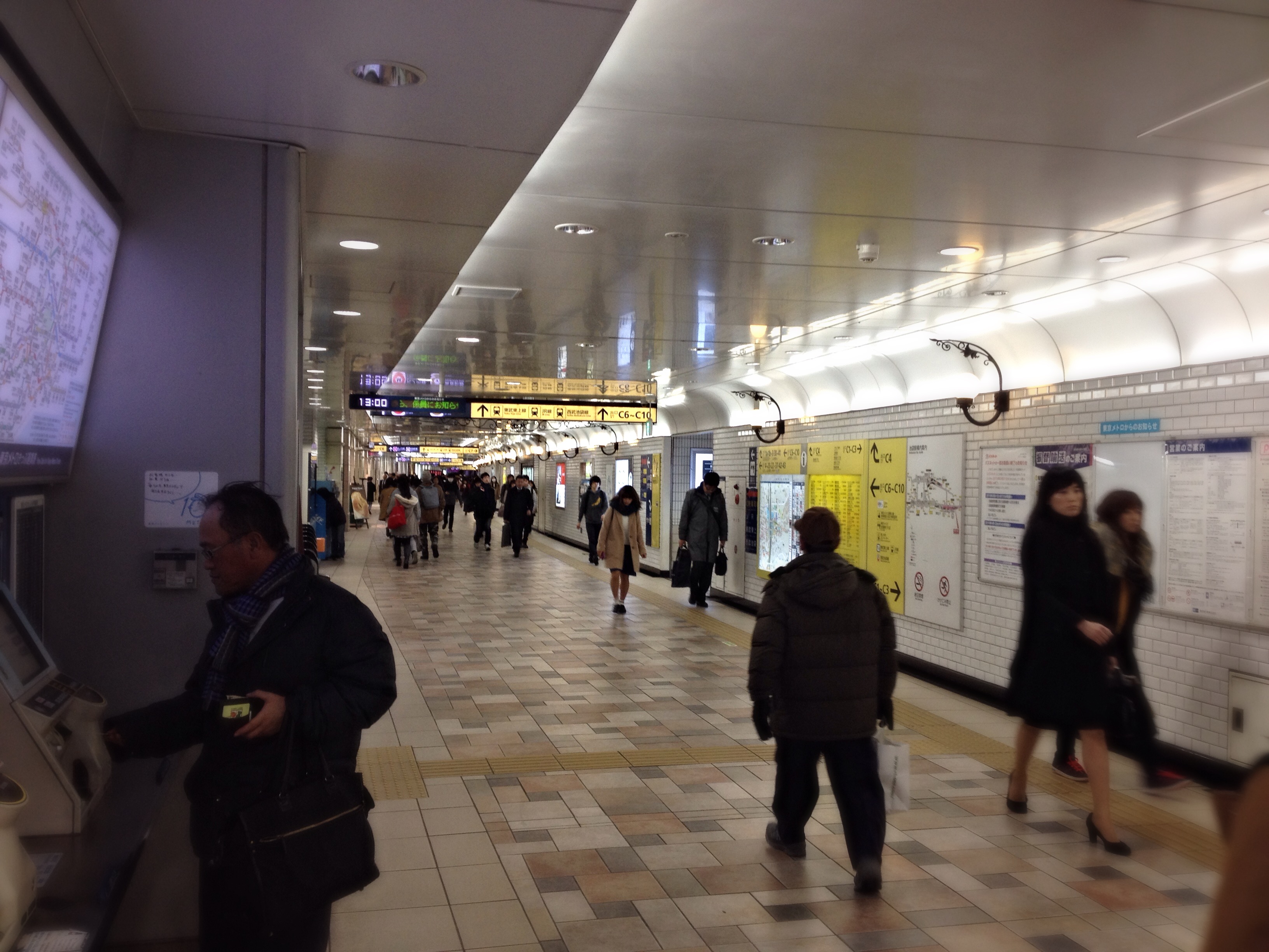 inside a train station in japan, blurry people walking by and a digital clock above shows 13:00