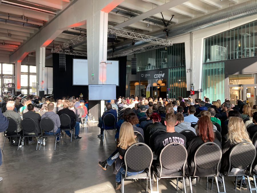 a hall with a bunch of people seen from behing who are seated on chairs and looking at a stage that is not visible on the photo, but you can guess because there is a big white screen above it