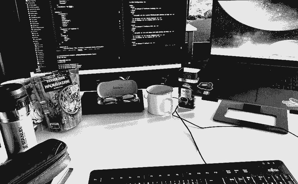 Black-and-white photo of a developer's workstation with two screens displaying code, a mechanical keyboard, a mouse, a coffee cup, and journal with pencil case. Black-and-white photo of a developer's workstation with two screens displaying code, a mechanical keyboard, a mouse, a coffee cup, and journal with pencil case.